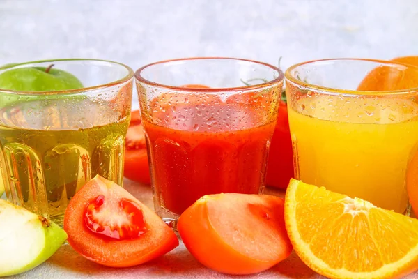 Vasos con naranja fresca, manzana, jugo de tomate sobre una mesa de hormigón gris. Lobulillos Frutas y verduras alrededor . — Foto de Stock