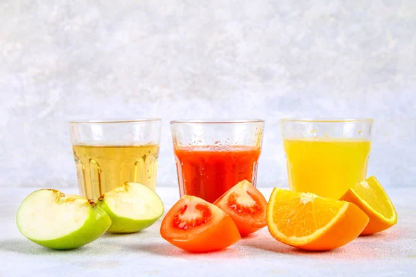Vasos con naranja fresca, manzana, jugo de tomate sobre una mesa de hormigón gris. Lobulillos Frutas y verduras alrededor . — Foto de Stock