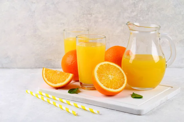 Glass cups and a pitcher of fresh orange juice with slices of orange and yellow tubes on a light gray table. — Stock Photo, Image