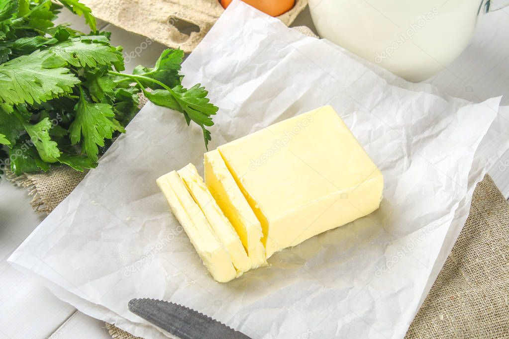 A bar of butter is cut into pieces on a wooden board with a knife, surrounded by milk, eggs and parsley on a white table. Ingredients for cooking.