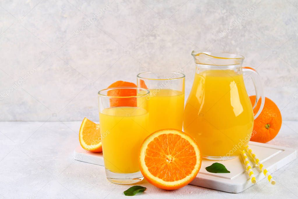 Glass cups and a pitcher of fresh orange juice with slices of orange and yellow tubes on a light gray table.