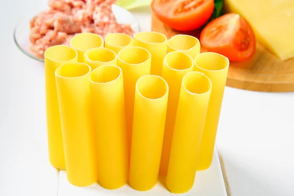 Cannelloni di pasta italiana. Tubo crudo per farcire ripieno circondato da ingredienti per la cottura, parmigiano, pomodoro, carne macinata su un tavolo di legno bianco . — Foto Stock