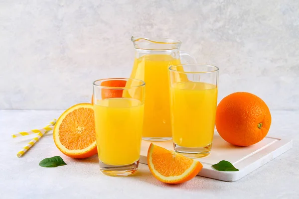 Glass cups and a pitcher of fresh orange juice with slices of orange and yellow tubes on a light gray table. — Stock Photo, Image