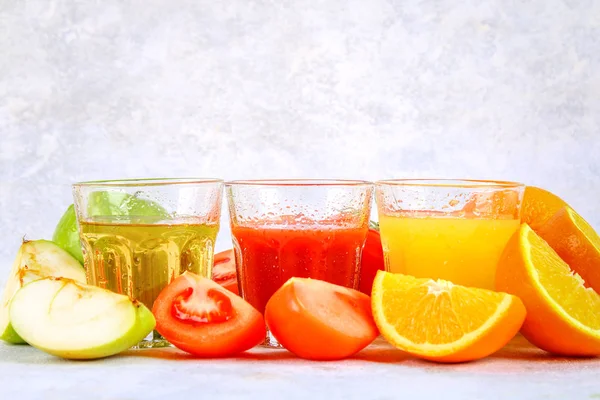 Vasos con naranja fresca, manzana, jugo de tomate sobre una mesa de hormigón gris. Lobulillos Frutas y verduras alrededor . — Foto de Stock