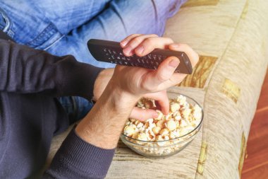 Bir adam bir kase patlamış mısır ve elinde bir uzaktan kumanda ile Tv kanepede görünüyor..