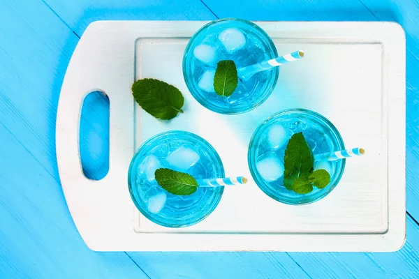 Blauer Cocktail mit Eis und Minze in Gläsern auf einem weißen Holzbrett auf einem blauen Tisch. Ansicht von oben. — Stockfoto