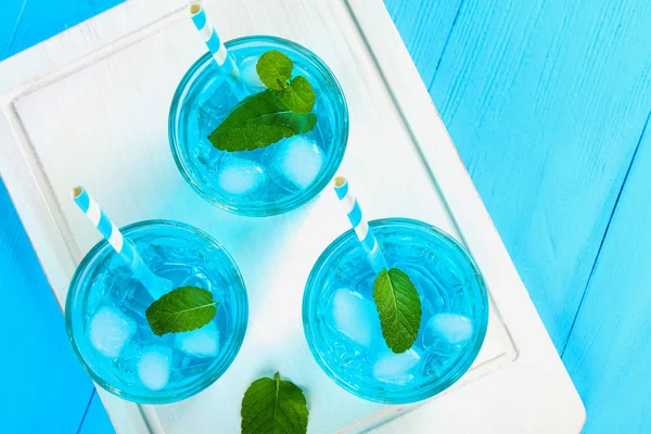 Blauer Cocktail mit Eis und Minze in Gläsern auf einem weißen Holzbrett auf einem blauen Tisch. Ansicht von oben. — Stockfoto