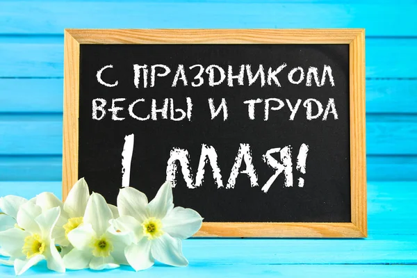 Chalkboard com o texto em russo: com as férias da primavera e do trabalho, 1 de maio. Flores brancas de narcisos em uma mesa de madeira azul . — Fotografia de Stock