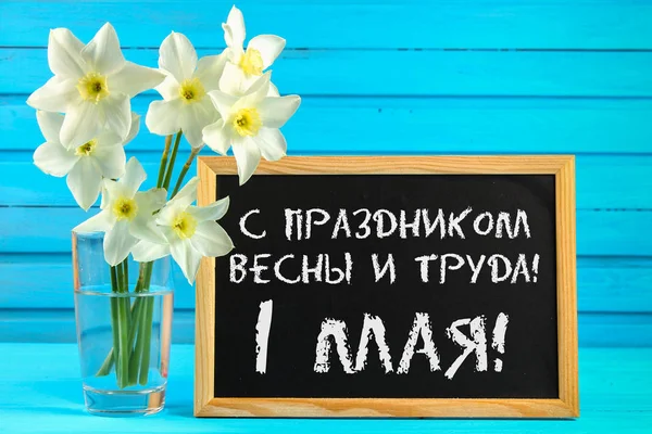 Lavagna con il testo in russo: con la festa della primavera e del lavoro, 1 maggio. Fiori bianchi di narcisi su un tavolo di legno blu . — Foto Stock