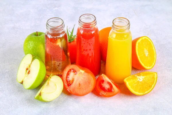 Bottles with fresh orange, apple, tomato juice on a gray concrete table. Fruits and vegetables around. — Stock Photo, Image