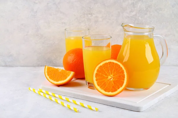 Glass cups and a pitcher of fresh orange juice with slices of orange and yellow tubes on a light gray table. — Stock Photo, Image
