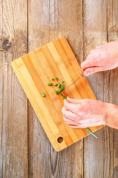 Αρσενικό χέρια κόβουμε ένα πράσινο κρεμμύδι σε ένα ξύλο κοπής σε ένα παλιό ξύλινο τραπέζι. — Φωτογραφία Αρχείου