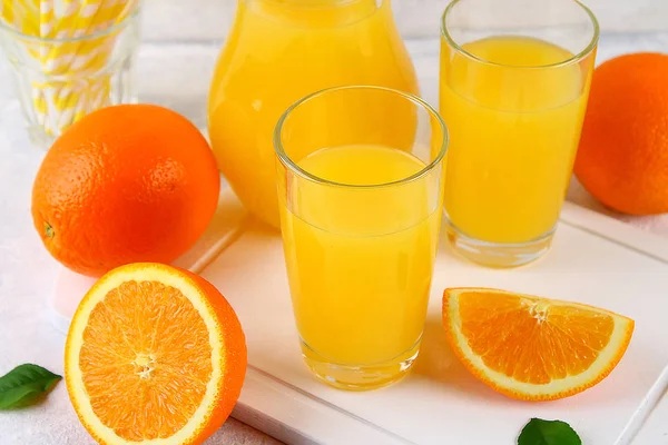 Bicchieri di vetro e una brocca di succo d'arancia fresco con fette di tubi arancioni e gialli su un tavolo grigio chiaro . — Foto Stock