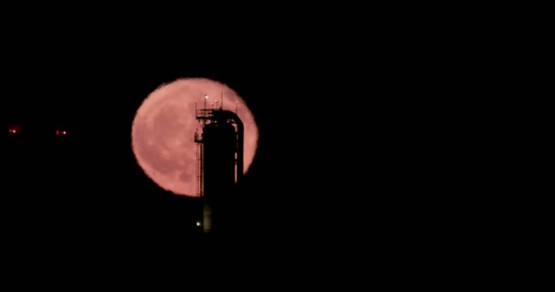 Red full moon rising from behind a large oil refinery — Stock Video