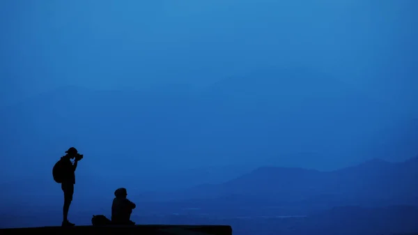 Homme debout et femme assise se relaxant au sommet d'une montagne et profitant d'une vallée. Belle vue sur le ciel panorama. Voyage style de vie randonnée concept vacances en plein air. Classic blue 2020 année concept de couleur — Photo