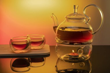 Cam tezgahta cam çaydanlık, sarı zemin üzerinde mum, cam çaydanlıkta siyah çay ve cam fincanlar ile ısıtılır.