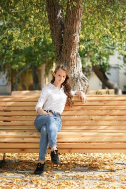  Sonbahar Park meydanında beyaz bluzlu ve kot pantolonlu bir kız ahşap bir bankta oturuyor, sonbaharı kaçırıyor.                              