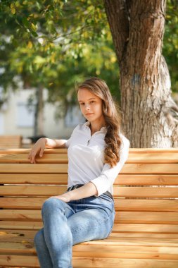  Sonbahar Park meydanında beyaz bluzlu ve kot pantolonlu bir kız ahşap bir bankta oturuyor, sonbaharı kaçırıyor.                              
