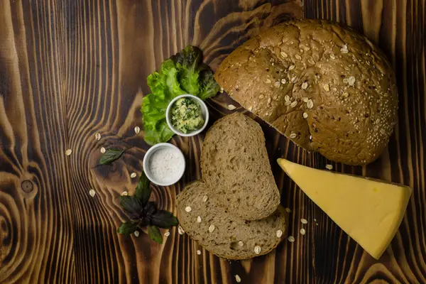 The bread with a piece of cheese, decorated with leaves of salad and basil with cups of butter are the wooden background — Stock Photo, Image