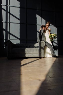 Gülümseyen gelin damat kanepe ve koluna oturan kucaklar. Onlar tarafından penceresinden sert ışık yanıyor