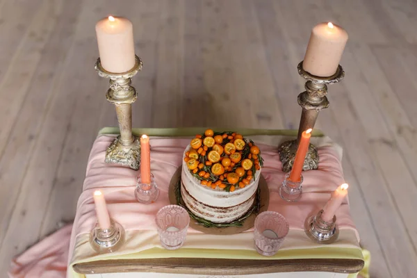 A wedding cake decorated with kumquat, sea buckthorn stands on a table with a pink cloth surrounded by candles. Top view