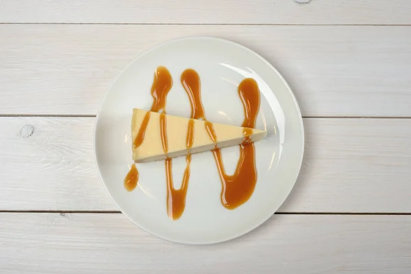 Un trozo de pastel de queso, rociado con salsa de caramelo en un plato blanco de pie sobre una mesa blanca de madera. La vista desde arriba — Foto de Stock