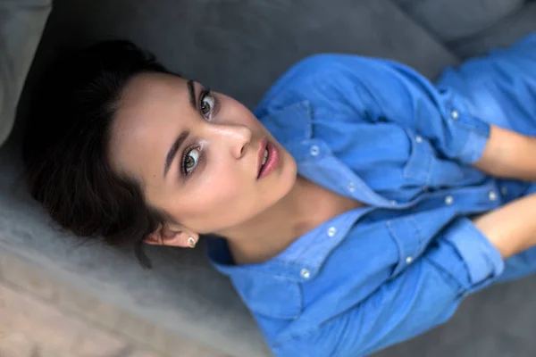 Retrato de uma jovem morena deitada em um sofá cinza em uma camisa jeans azul — Fotografia de Stock