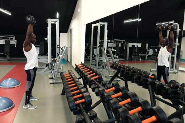 Muscular black man in a white t-shirt doing exercises with dumbbells in front of the mirror — Stock Photo, Image