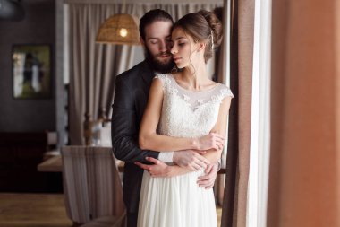 Gelin ve damat bir odada cam kenarında duruyor. Damat gelini arkadan sarılma
