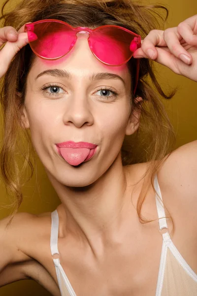 Retrato de uma jovem com levantamentos capilares coletados óculos cor de rosa na testa e mostrando a língua em um fundo amarelo — Fotografia de Stock