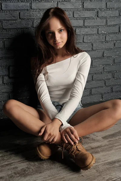 Young brunette in white shirt, shorts and boots sitting on the floor cross-legged and leaning against the brick wall — Stock Photo, Image