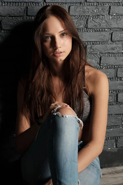Jonge brunette in crop top en gescheurde spijkerbroek, zittend op de vloer leunend tegen de muur — Stockfoto
