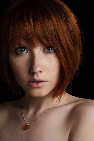 Retrato de una joven con el pelo rojo y pecas con un colgante en el cuello en forma de corazón mirando a la cámara sobre fondo oscuro — Foto de Stock