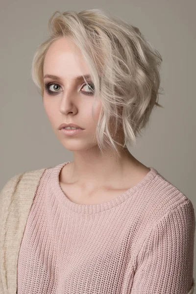 Retrato de chica de rubia con maquillaje de ojos oscuros y pelo corto, tirado por un lado, suéter rosa claro sobre un fondo claro —  Fotos de Stock