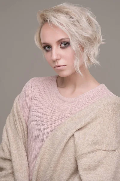 Retrato de chica de rubia con maquillaje de ojos oscuros y pelo corto en un suéter de color rosa pálido y chaqueta beige de gran tamaño, mirando a la cámara sobre fondo claro — Foto de Stock