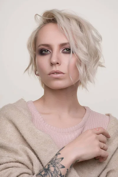 Portrét dívky Blondýna s tmavé oční make-up a krátké vlasy ve světle růžový svetr stojí na světlém pozadí pohledu do kamery, Foto níže — Stock fotografie