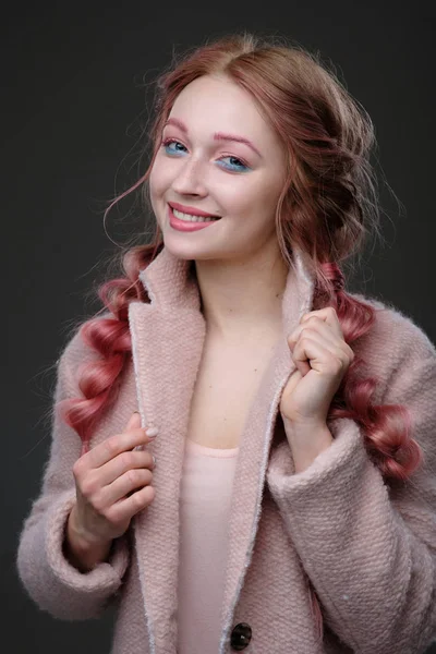 The girl with pink hair in braids, with pink-blue makeup with a pink coat, smiling, looking at the camera on gray background