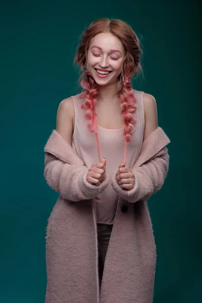 Η κοπέλα με ροζ μαλλιά σε πλεξούδες και ροζ-μπλε μακιγιάζ με ένα ροζ παλτό στέκεται σε σκούρο φόντο, κρατώντας τα άκρα των πλεξούδες με κλειστά τα μάτια και χαμογελά — Φωτογραφία Αρχείου