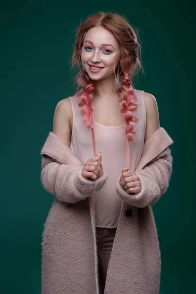 Η κοπέλα με ροζ μαλλιά σε πλεξούδες και ροζ-μπλε μακιγιάζ με ένα ροζ παλτό στέκεται σε σκούρο φόντο, κρατώντας τις άκρες από τα κοτσιδάκια βλέπουν φωτογραφική μηχανή και χαμογελαστός — Φωτογραφία Αρχείου
