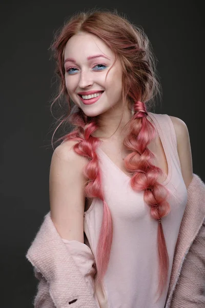 The girl with pink hair in braids, and pink-blue makeup with a pink coat worn off the shoulder stands in half-turn on a dark background and looking at camera, smiling