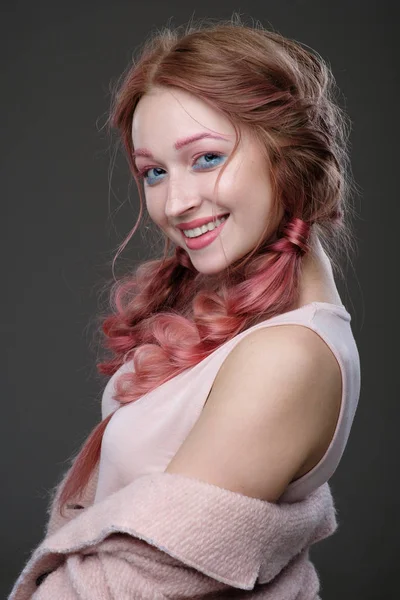 Close-up portret van het meisje met roze haren in vlechten, en roze-blauw make-up met een roze jas versleten uit de schouder permanent in de half-bocht op een donkere achtergrond glimlachen en kijken naar camera — Stockfoto