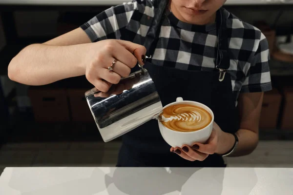 Barista girl cream draws art on the cappuccino with the coffee up and small jug in her hands