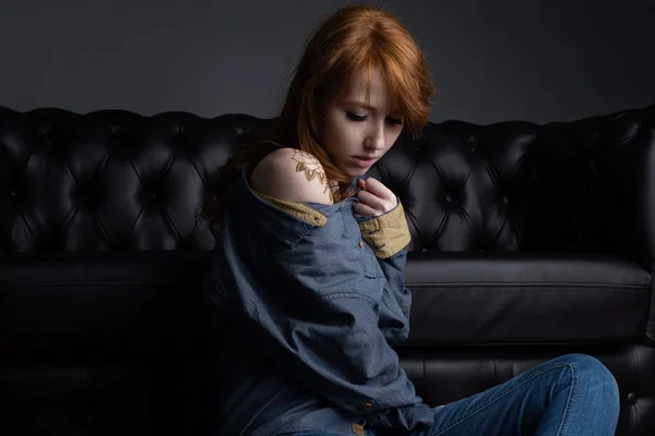 Retrato de uma menina ruiva com uma camisa jeans sentada no sofá preto e olhando para baixo . — Fotografia de Stock