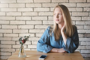 Genç sarışın mavi gömlekli boyun yakınındaki bir sol el yapımı bir kafede masada oturuyor. Bir telefon, bir tuğla duvar zemin karşı geçerli.