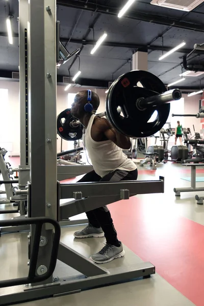 A black muscular guy in a white tank top, in blue headphones squats with a barbell. — Stock Photo, Image