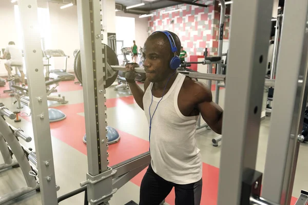 A black muscular guy in a white T-shirt, wearing blue headphones stands with a barbell on his shoulders. View from above. — Stock Photo, Image