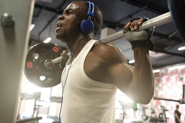 Un tipo musculoso negro con una camiseta blanca en auriculares azules sostiene una barra en sus hombros . — Foto de Stock