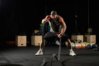 Spor üniformalı kaslı bir adam ve beyzbol şapkası askıda iplerle vahşice egzersiz yapıyor.