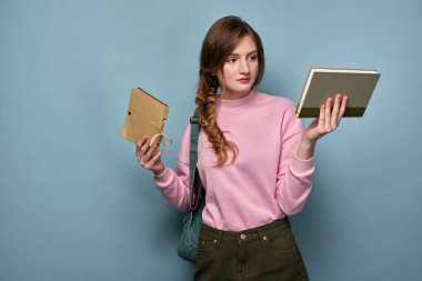 Pembe kazaklı mavi gözlü bir kız elinde kitaplarla mavi arka planda duruyor..