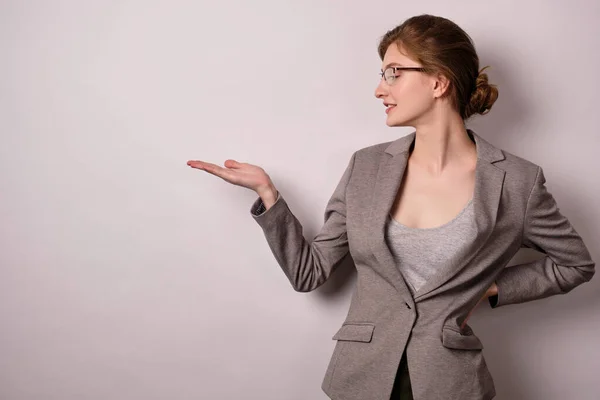 Flicka med glasögon och en grå jacka, stående på en ljus bakgrund vrider huvudet åt sidan och pekar på det tomma utrymmet. — Stockfoto